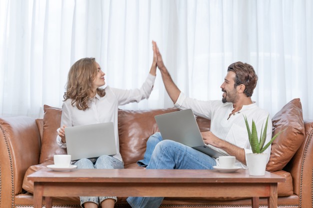 alt= "Casal sentado no sofá, com um notebook cada um, comemorando"